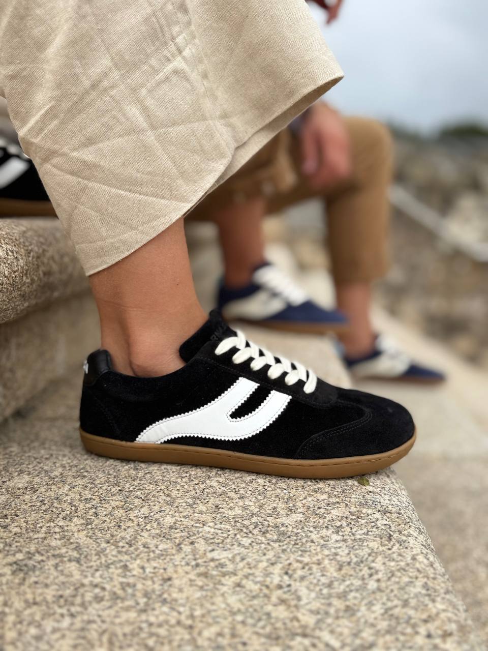 Persona sentada en escalones de piedra con zapatillas barefoot Atlantic Coal negras, detalle lateral — ARNASA